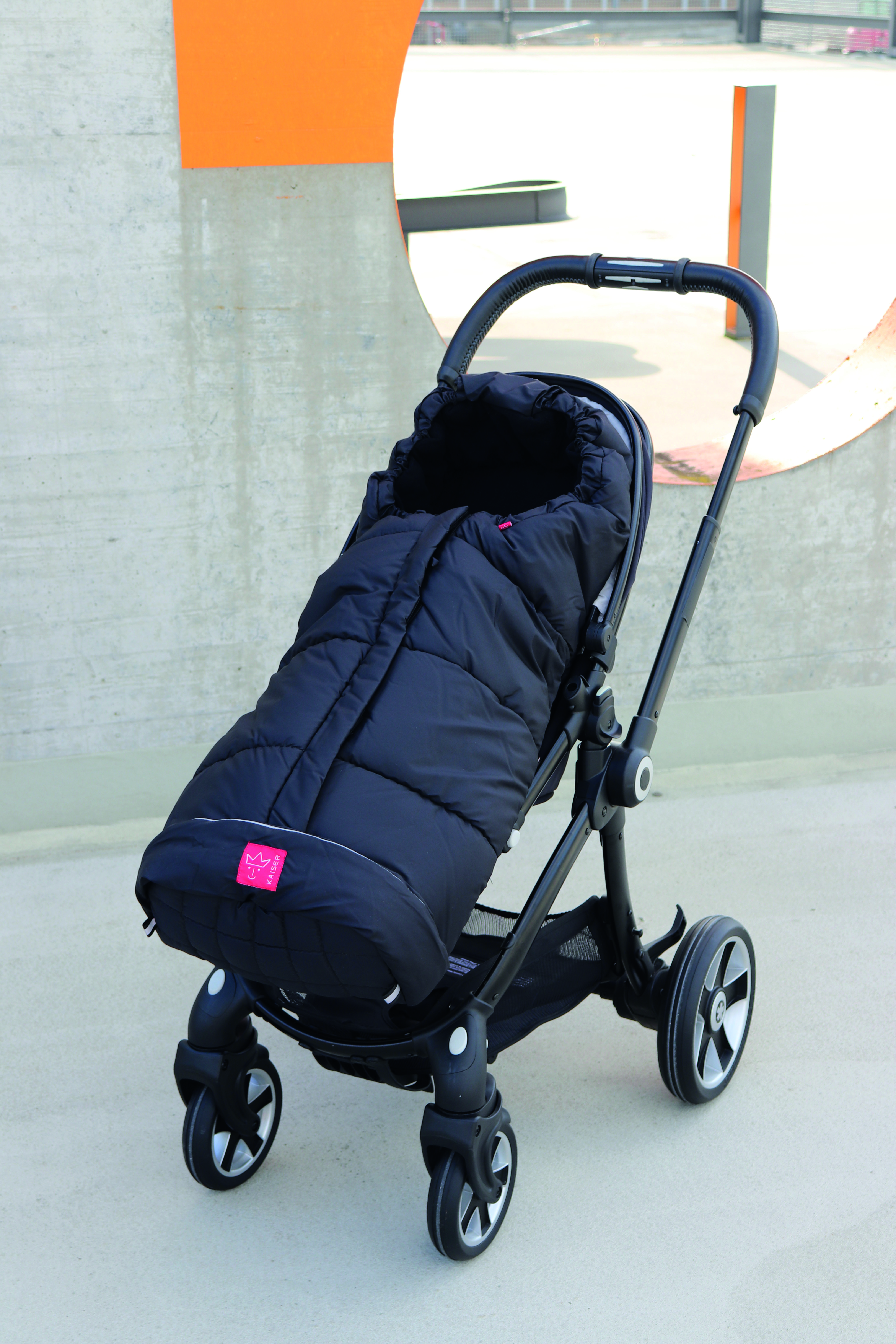 Schwarzer Fußsack im Kinderwagen montiert – Außenansicht Fußsack in Schwarz in einem modernen Kinderwagen montiert, Vorderansicht, draußen vor Betonwand fotografiert.